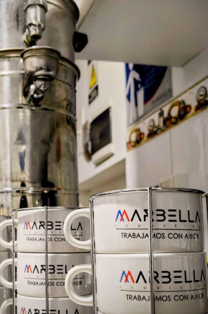 Tazas de café con el logo "Marbella Coworking - Trabajamos con Amor" apiladas junto a una cafetera, simbolizando el café ilimitado como beneficio para coworkers en Bogotá.