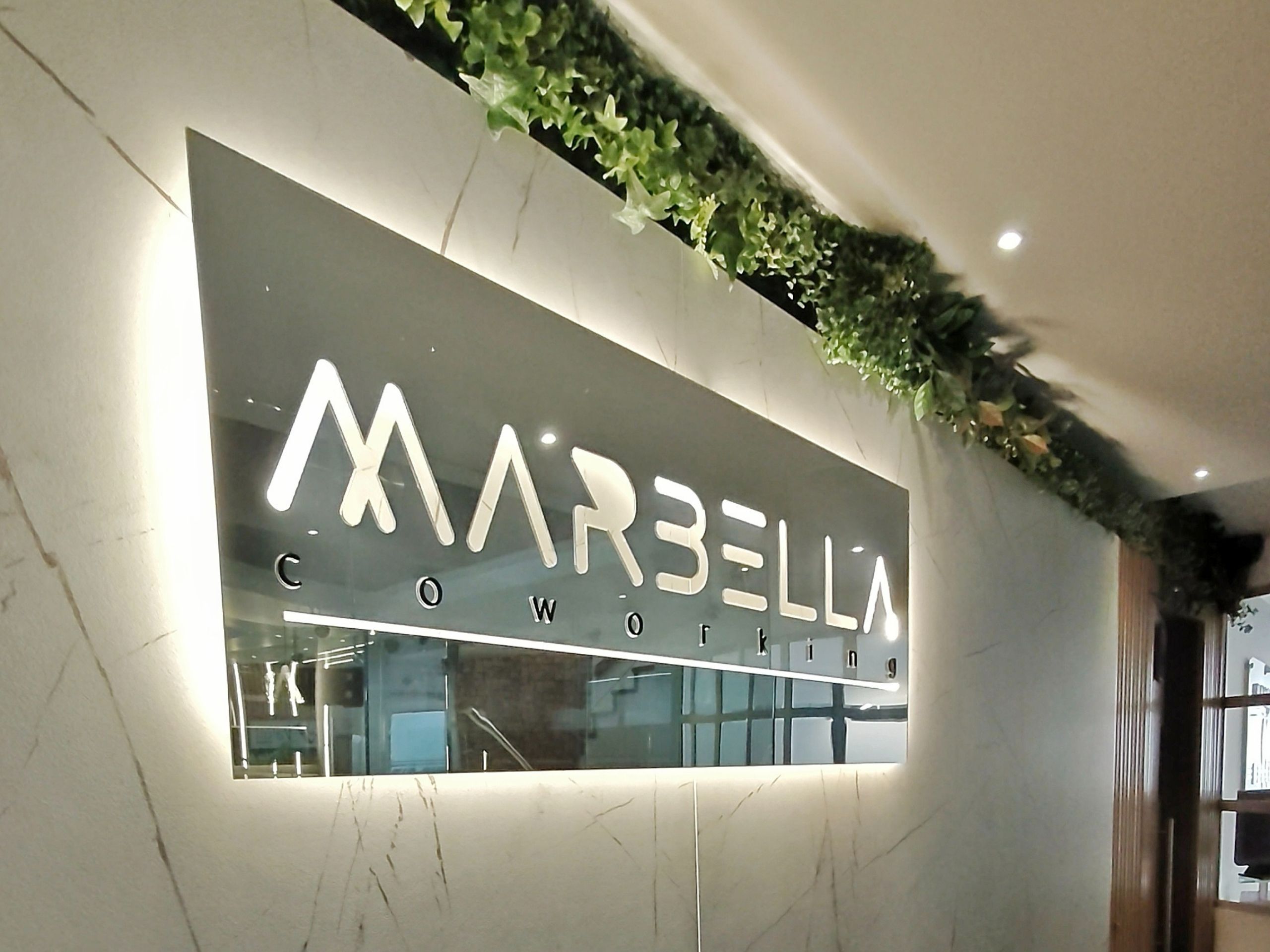Imponente logo iluminado de Coworking Marbella en la recepción, con detalles de vegetación, simbolizando una cálida bienvenida y la conexión con nuestra marca en Bogotá.