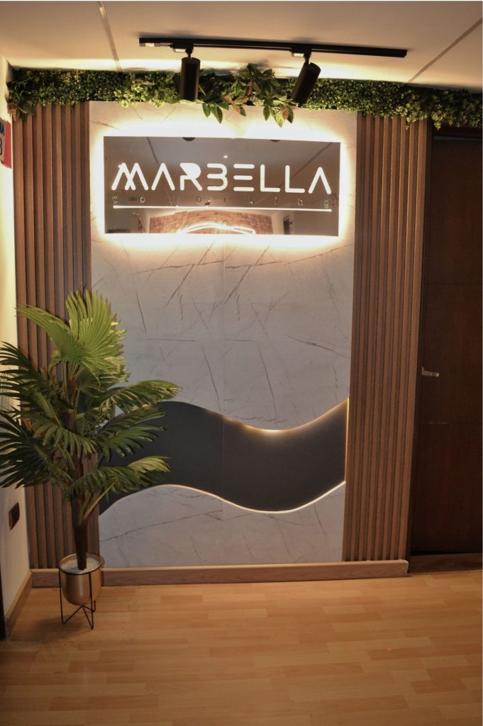 Logo iluminado de Coworking Marbella en una pared con diseño moderno de madera y mármol, con plantas decorativas, reflejando un ambiente profesional y acogedor.