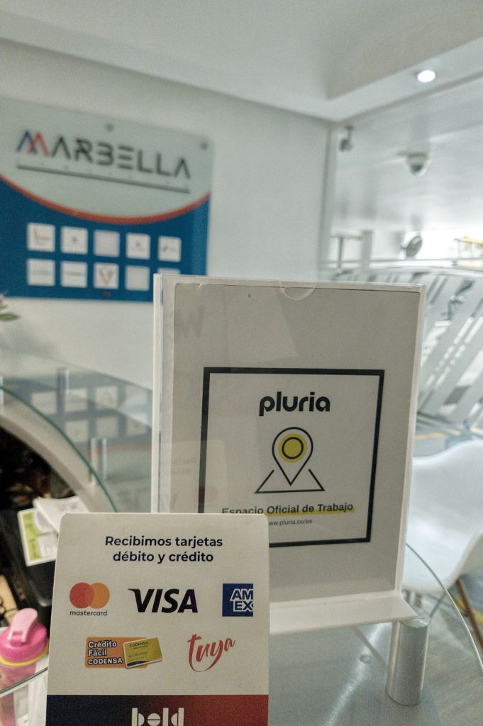 Punto de pago en la recepción de Coworking Marbella, mostrando aceptación de tarjetas débito/crédito (Visa, Mastercard, Amex) y el logo de Pluria, facilitando el acceso a nuestros espacios WorkOffice en Bogotá.