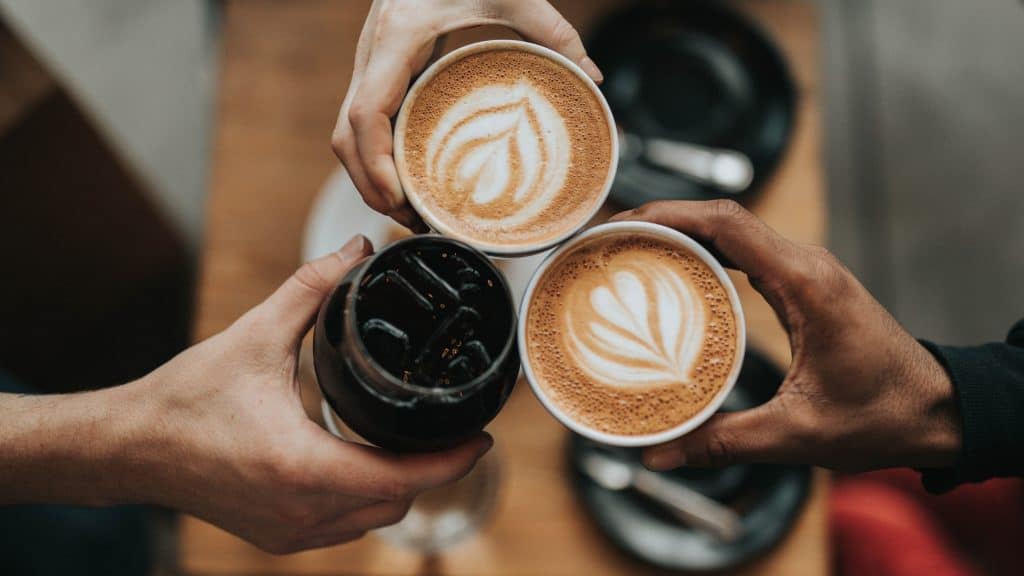 Tres personas brindando con diferentes tipos de café: un latte art, un café negro y un latte art en una taza de papel. La imagen representa la cultura del café y la conexión social en un ambiente de coworking.