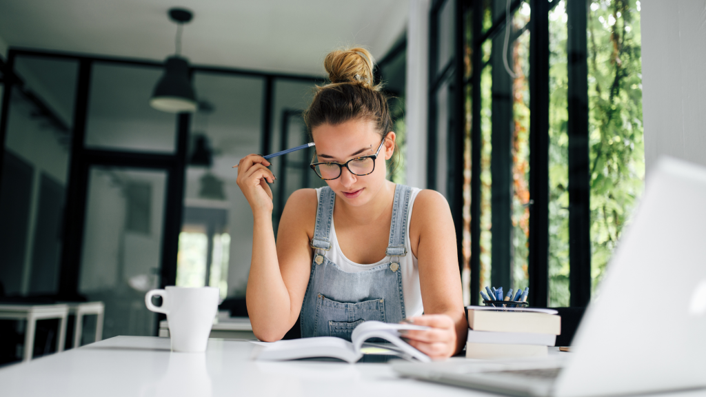 Una mujer joven, con un moño y gafas, se sienta en un escritorio de oficina con un libro y una laptop, sosteniendo un lápiz en su mano. Su expresión pensativa indica enfoque y concentración en un ambiente de trabajo.